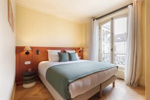 A hotel room with a large bed, a nightstand, a window with a view of buildings, and a balcony with a railing. - A Charming 1-BR with Services in Opera-St-Lazare (Paris)