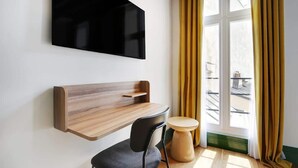 Una habitación moderna con una televisión montada, un estante de madera, una mesa redonda, una silla y una ventana con cortinas.