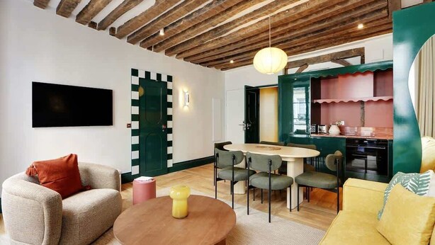 Una sala moderna con techo de madera, un televisor de pantalla plana, una mesa de comedor redonda con sillas y una cocina con una pared verde.