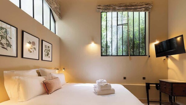 A hotel room with a large bed, a desk, a flat-screen TV, and a window with vertical blinds. - A Charming 2-BR/2Ba in Saint Germain (Paris)