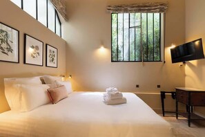 A hotel room with a large bed, a desk, a flat-screen TV, and a window with vertical blinds. - A Charming 2-BR/2Ba in Saint Germain (Paris)