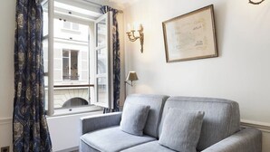 A room with a grey sofa, patterned curtains, a framed picture, and a window with shutters. - French Chic Apartment in Saint Germain #1 (Paris)