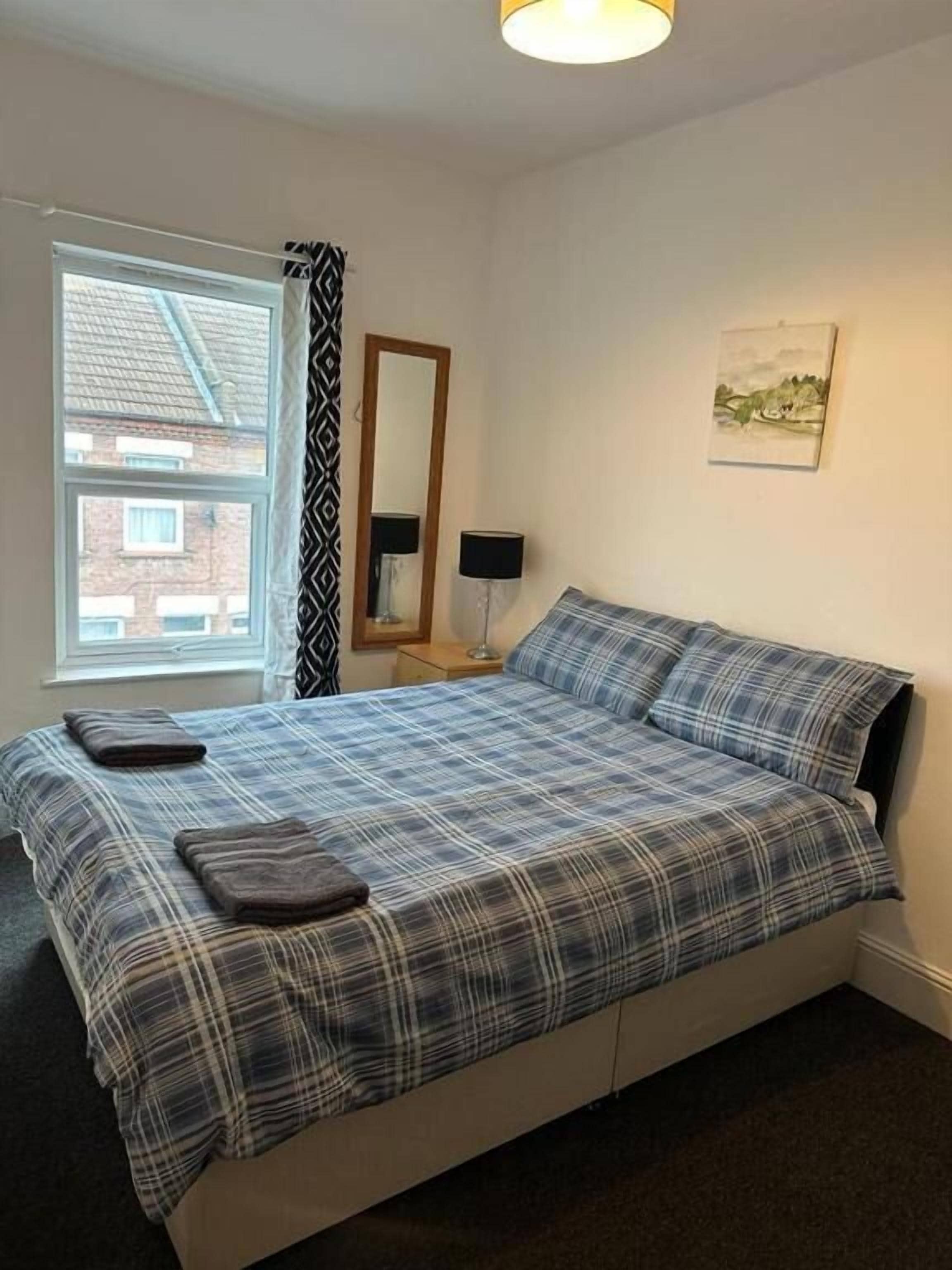 A bedroom with a bed, a window, a mirror, a lamp, and a painting on the wall.