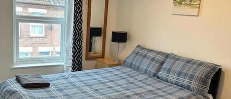 A bedroom with a bed, a window, a mirror, a lamp, and a painting on the wall.