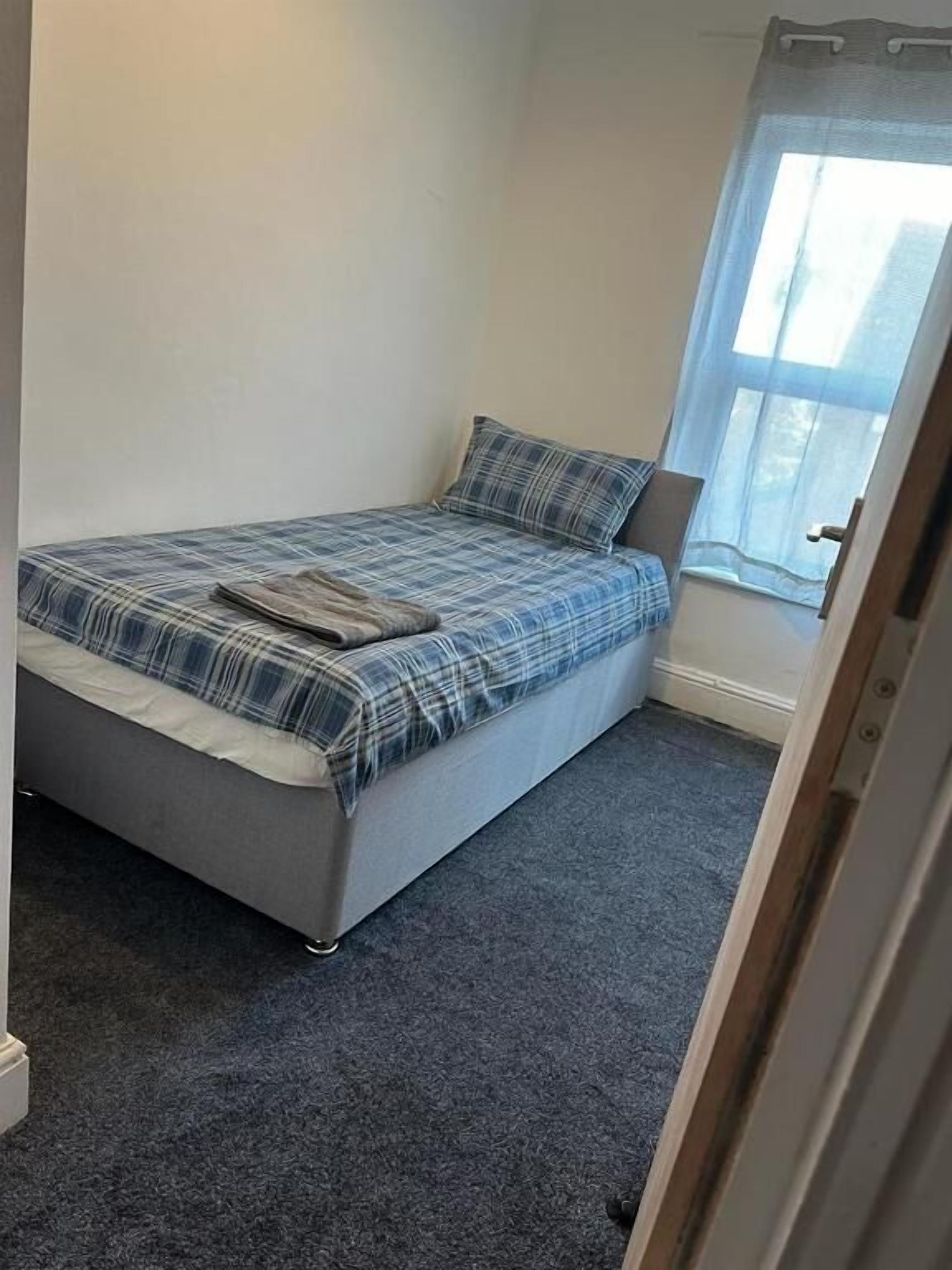 Una habitación pequeña con una cama individual, alfombra gris, colcha escocesa y una ventana con cortina translúcida.