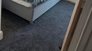 A small, single-bed room with a grey carpet, a plaid bedspread, and a window with a sheer curtain. - Luton Homestay near AIRPORT  (Luton)
