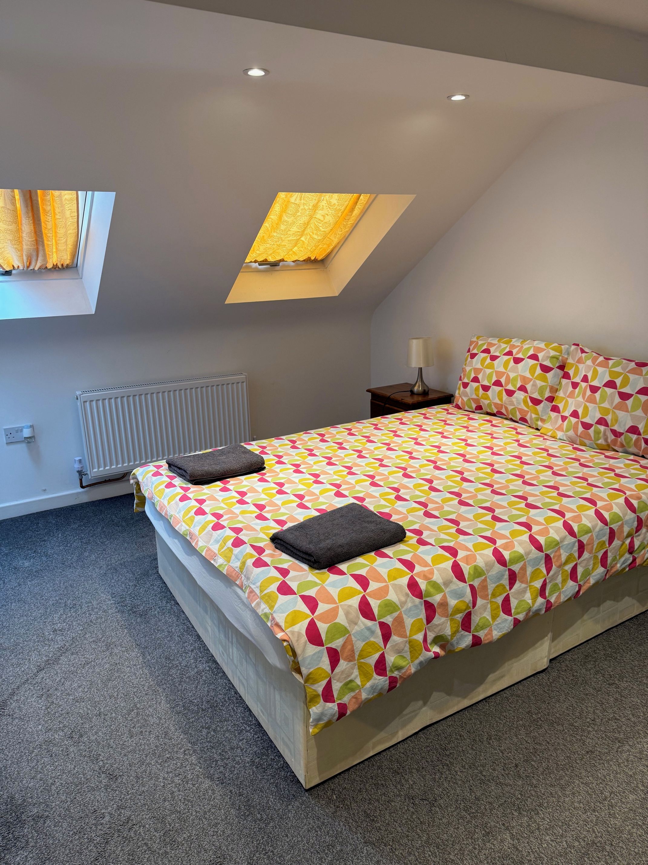 A bedroom with a bed, a nightstand, a lamp, a radiator, and a skylight.