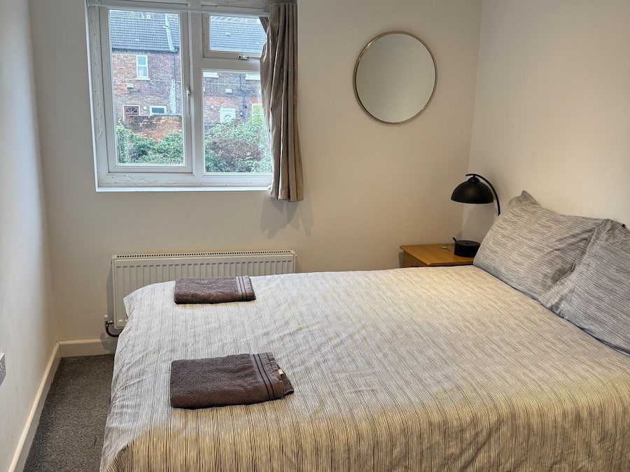 A bedroom with a bed, a window with curtains, a radiator, a bedside table with a lamp, and a view of a brick building outside.