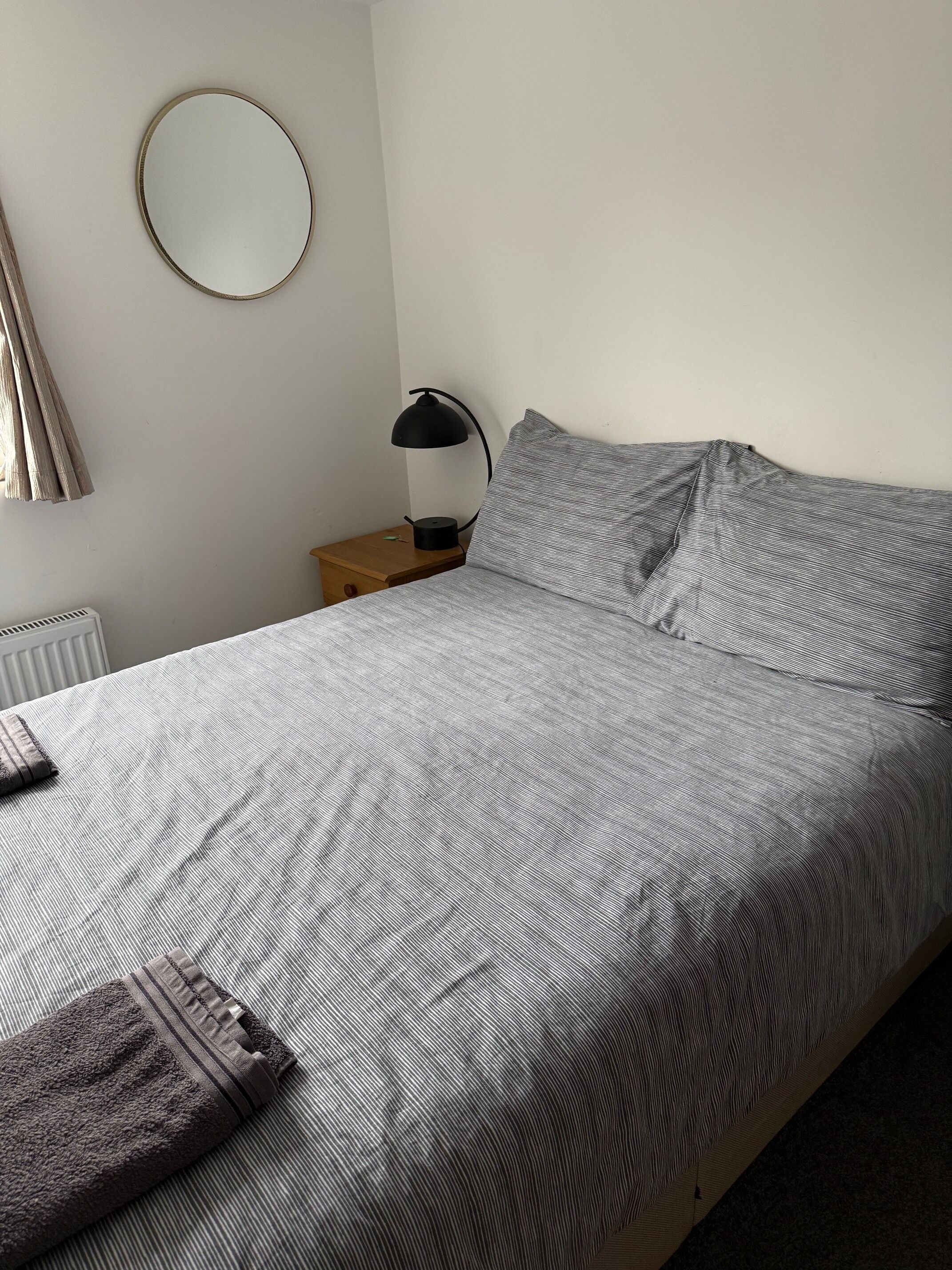 A neatly made bed with a striped bedspread, a round mirror on the wall, and a small bedside table with a lamp.