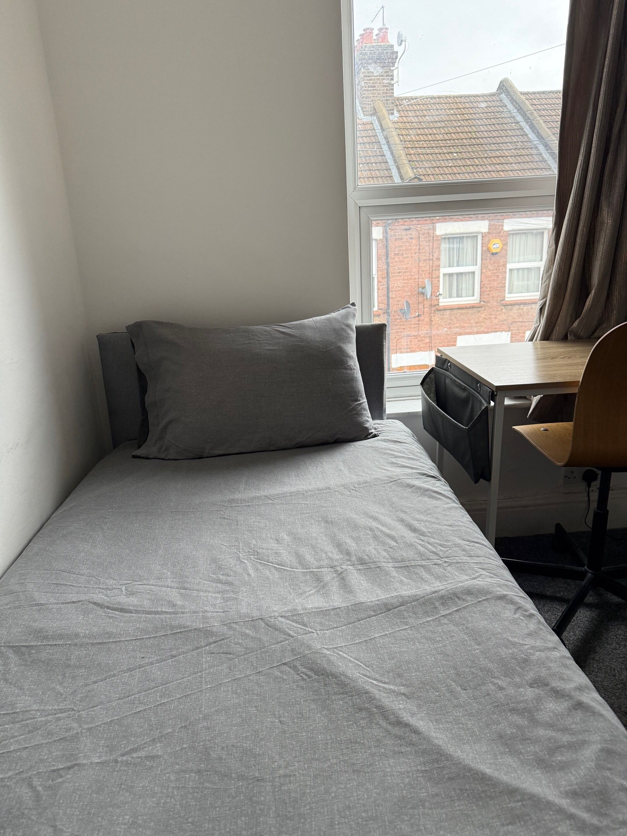 Una habitación pequeña con cama individual, una colcha gris, un escritorio de madera con silla y una ventana con cortinas.