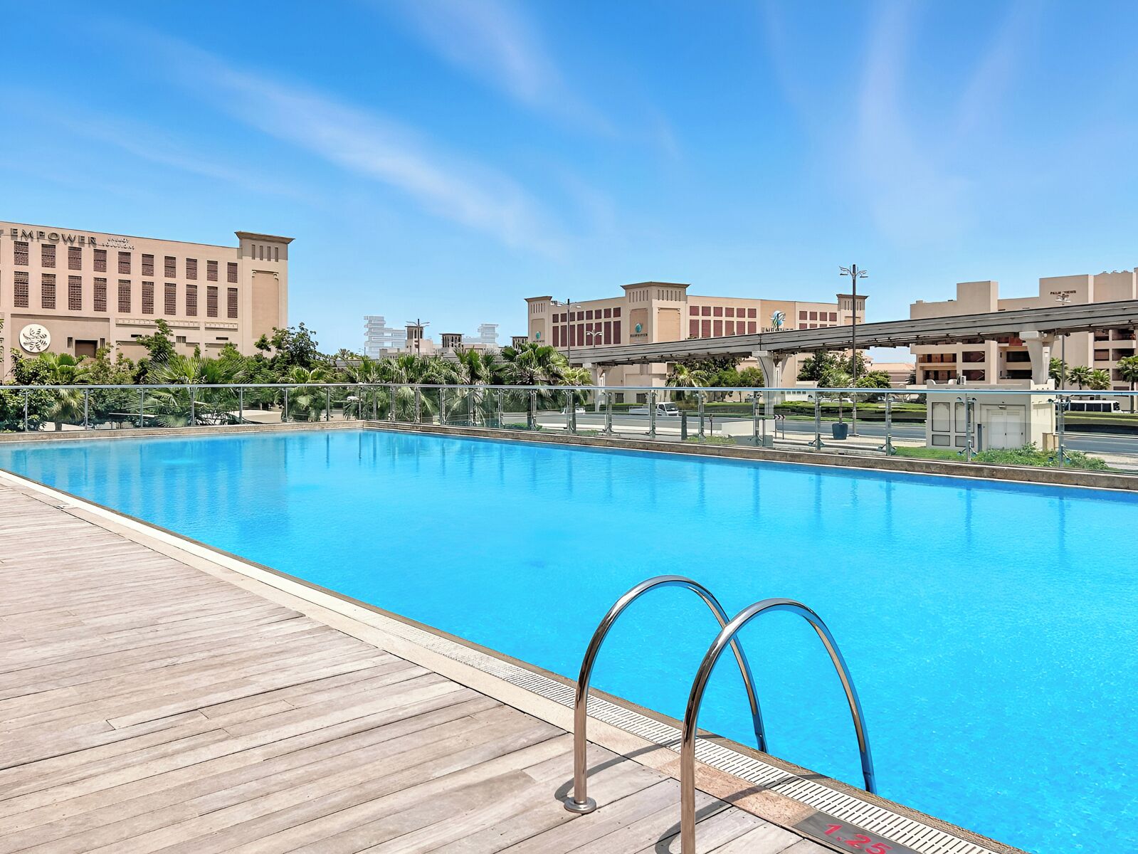 Una piscina con muelle de madera y escaleras metálicas, rodeada de edificios y palmeras.