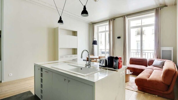 A modern kitchen with a central island, a dining area with a table and chairs, and a living area with a sofa and a TV. - A Bright 1-BR in Grands Boulevards (Paris)