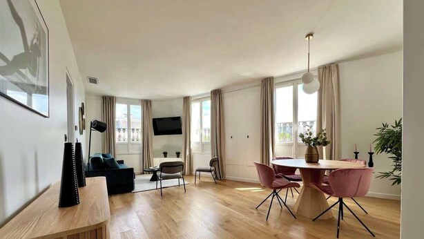 Ein geräumiges Wohnzimmer mit Holzboden, einem Essbereich mit rundem Tisch und rosa Stühlen, einem schwarzen Sofa und einem an der Wand befestigten Fernseher.