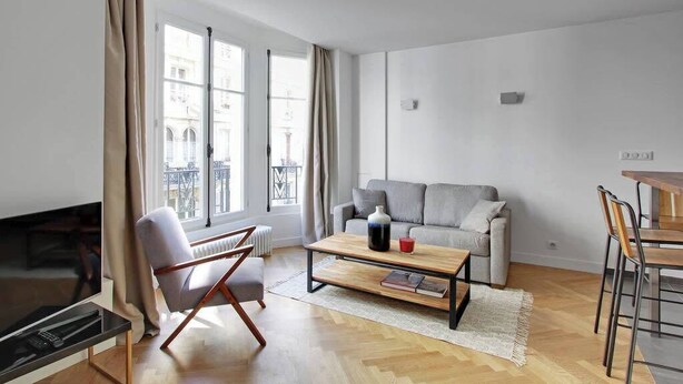 Una sala de estar moderna con un sofá gris, una mesa de centro de madera, un televisor y un comedor con una mesa y sillas.