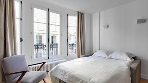 Un dormitorio con cama, una silla y ventanas amplias.