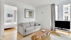 Ein modernes Wohnzimmer mit einem grauen Sofa, einem Holztisch, einem Flachbildfernseher und einem großen Fenster mit Vorhängen.