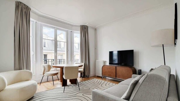 Ein modernes Wohnzimmer mit einer Couch, einem Esstisch mit Stühlen, einem Fernseher auf einem Schrank und großen Fenstern mit Vorhängen.