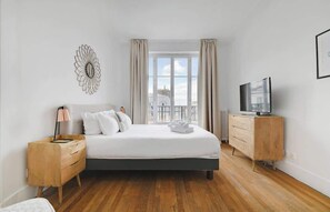 Ein Schlafzimmer mit einem Bett, Holznachttischen, einem Fernseher und Blick auf die Stadt durch große Fenster.