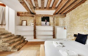 A bedroom with a stone wall, wooden beams, a bed with white linens, and a staircase. - A Stunning 1-BR Apartment close to the Louvre (Paris)
