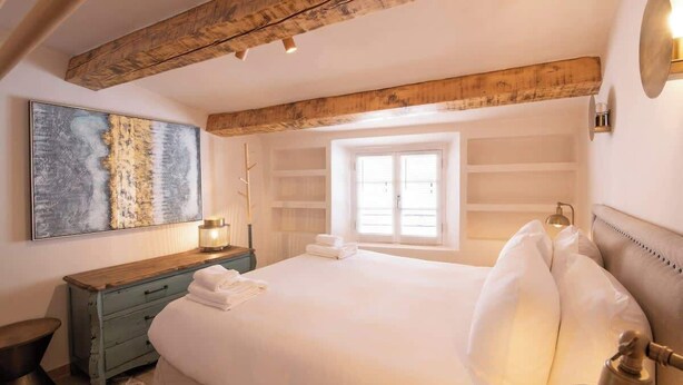 Une chambre avec un grand lit, des poutres en bois, une fenêtre, une table de chevet avec une lampe et un tableau au mur.