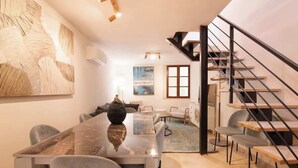 Un salon moderne avec une table à manger en marbre, des chaises recouvertes d’un tissu gris, un grand tableau abstrait, un escalier avec des marches en bois et une vue sur l’extérieur par une fenêtre.