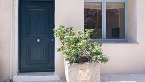 Eine dunkelblaue Tür mit einem kleinen Fenster darüber, einer Pflanze im Vorbereich und einem Fenster mit Jalousien zur Rechten.