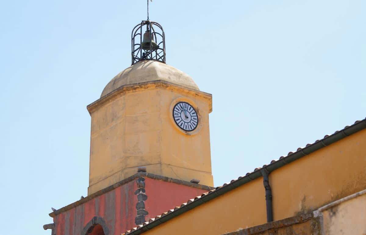 A clock tower with a bell on top, part of a building with a yellow facade and red roof.