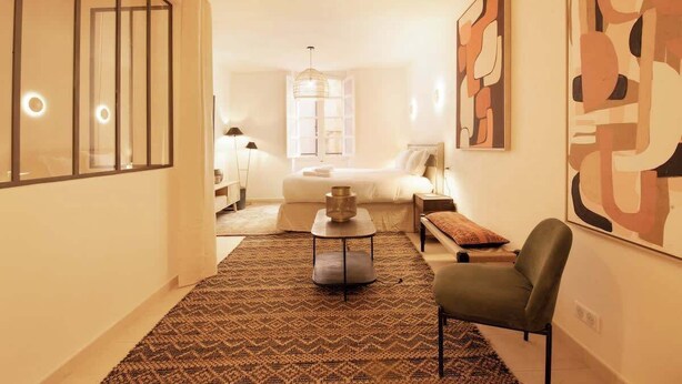 Une chambre d’hôtel moderne comprenant un lit, un coin salon avec un fauteuil et une table, un grand tableau abstrait et un tapis à motifs.