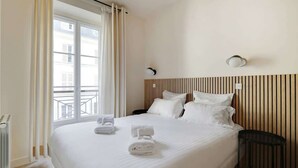 Hotellihuone, jossa on suuri sänky, valkoiset vuodevaatteet, puinen pääty, yöpöytä, seinään kiinnitetty lamppu ja ikkuna, jossa on verhot.