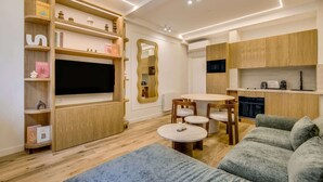 A modern living room with a flat-screen TV, wooden shelving, a dining area with a round table and chairs, and a kitchenette with wooden cabinets. - A Sublim 1-BR Apartment - Saint Germain (Paris)