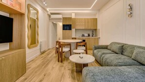 A modern living room with a grey sofa, wooden dining table, and chairs, a TV, and a kitchen area with wooden cabinets and a microwave. - A Sublim 1-BR Apartment - Saint Germain (Paris)