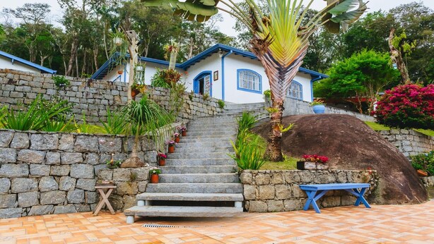 A house with a stone staircase leading to a blue door, surrounded by lush greenery and a large rock. - Country house with heated pool near São Paulo. (Mairiporã)