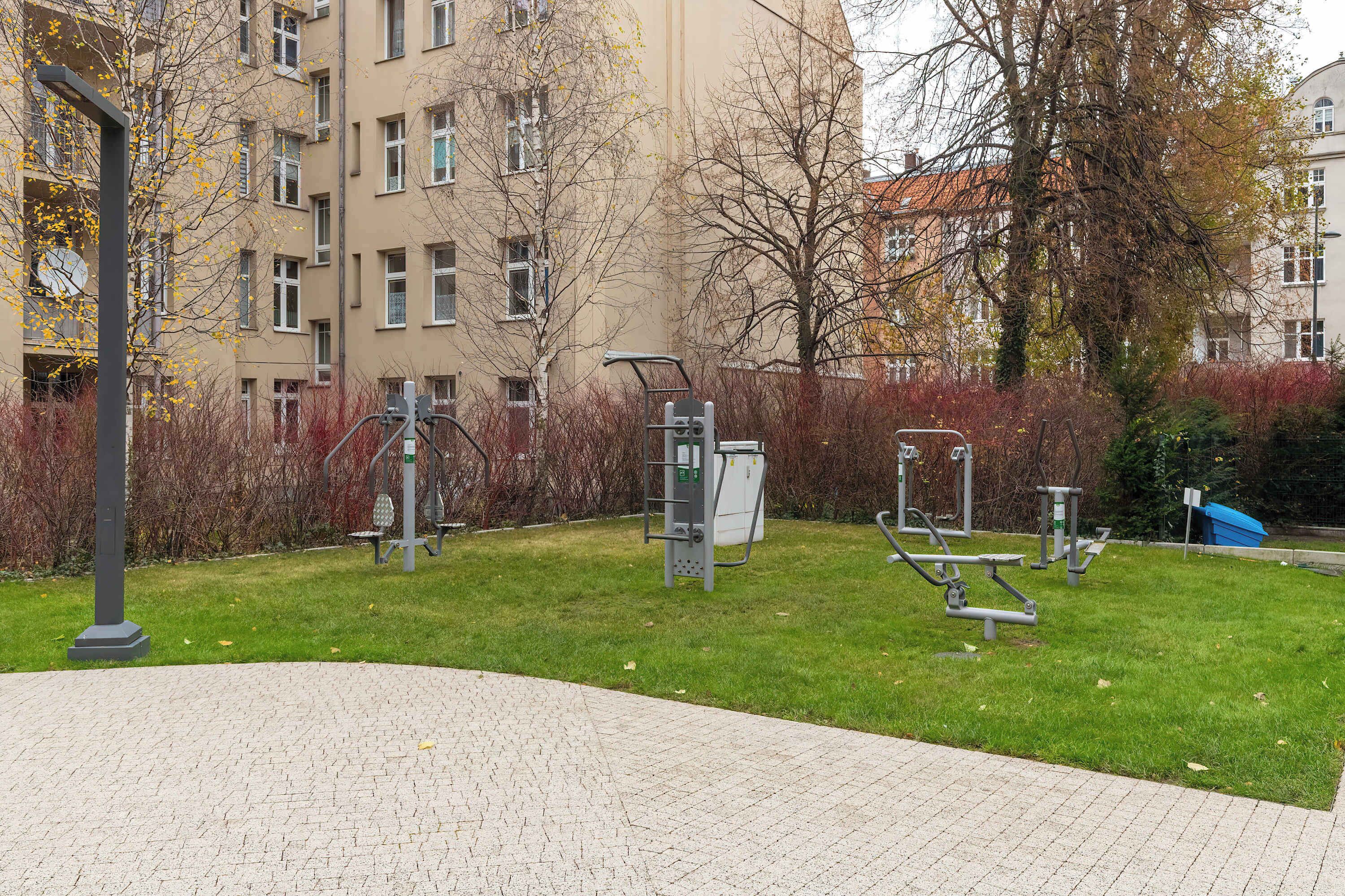 An outdoor gym area with exercise equipment, a paved walkway, and a building in the background.