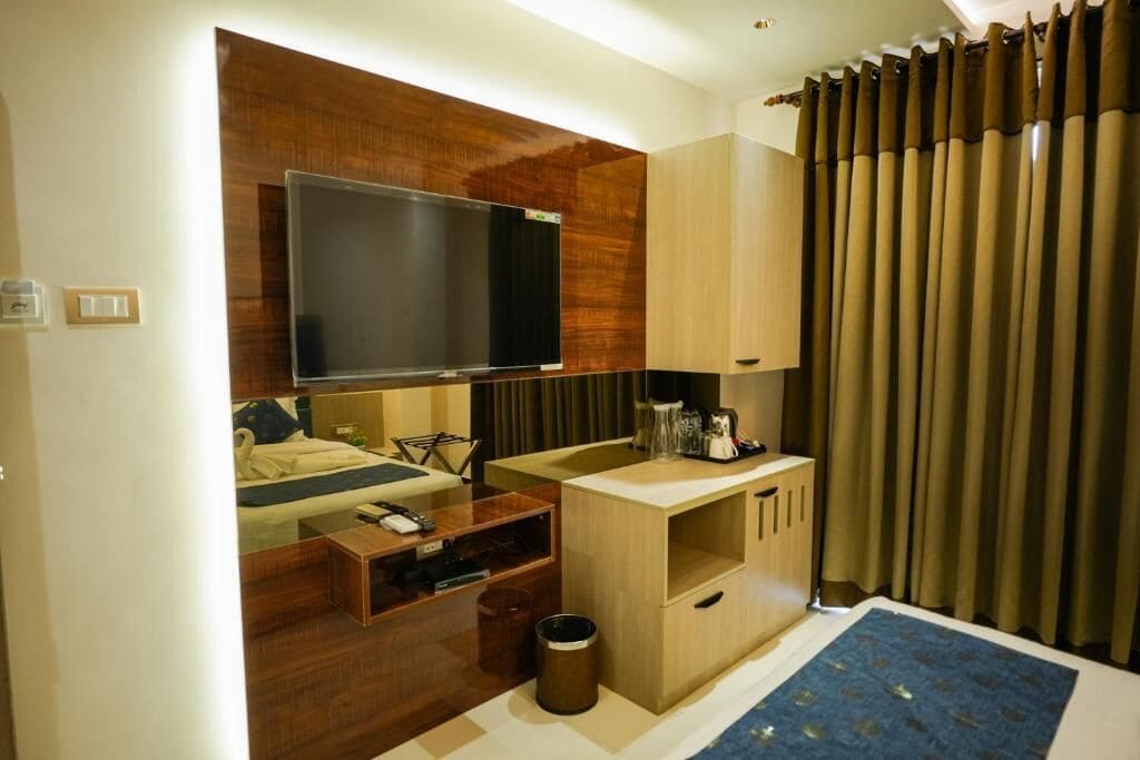 A modern hotel room with a flat-screen TV, wooden furniture, a small kitchenette, and a large window with curtains.