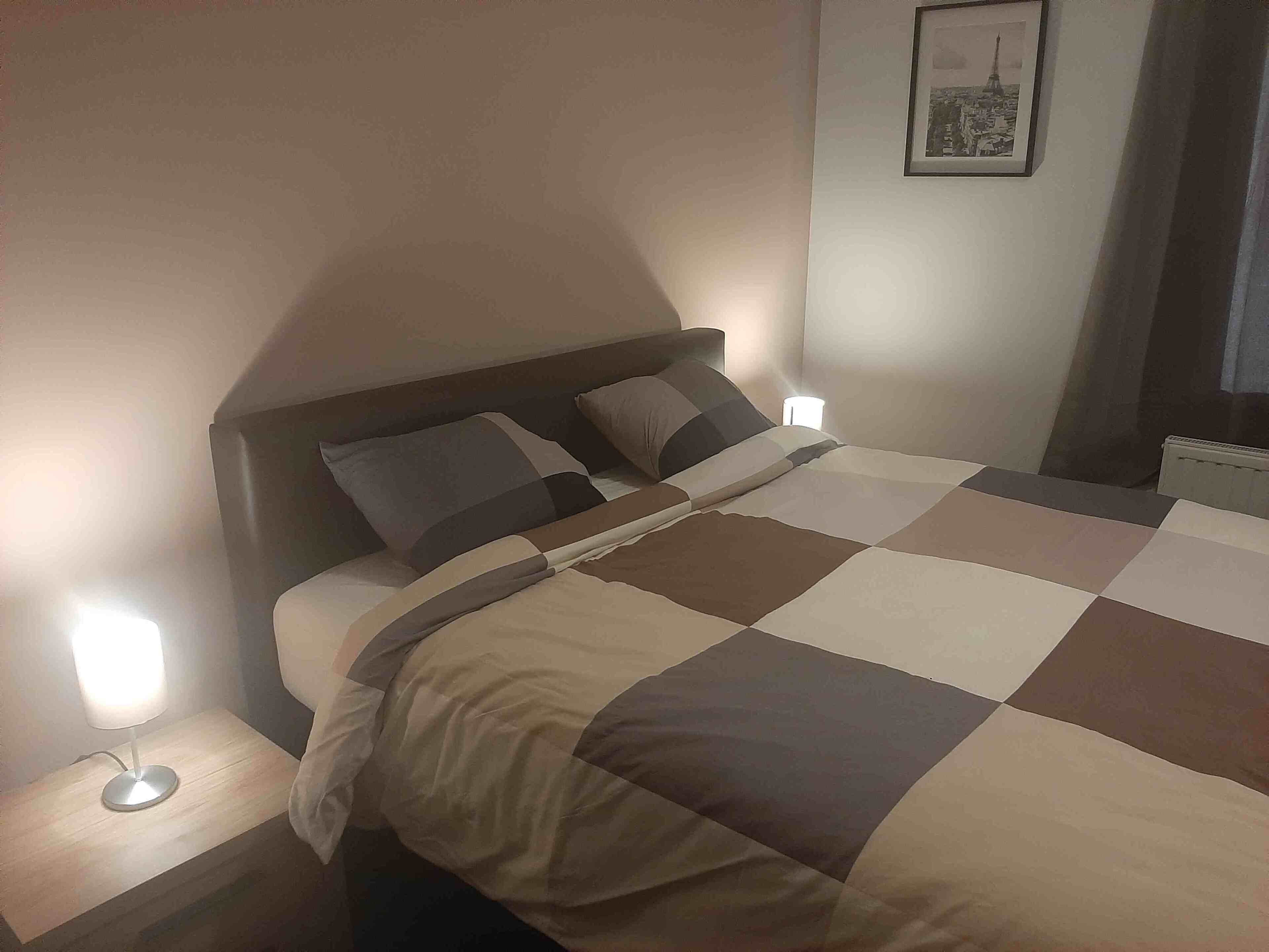 Un llit ben arropat amb un estovat a quadres, una làmpada de nit i un quadre al mur.