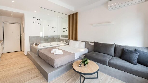 A modern living room with a sofa, a coffee table, and a bed with a glass wall. - Porto Sea View - Luxury Junior Suites (Thessaloniki)