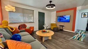 Un salon moderne avec un canapé, un fauteuil et une table basse ronde. Un téléviseur est fixé au mur, et une porte est visible au fond.