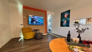 Un salon moderne avec un plancher en bois, un fauteuil jaune, un téléviseur, une bouteille de vin et un plateau de fromages.