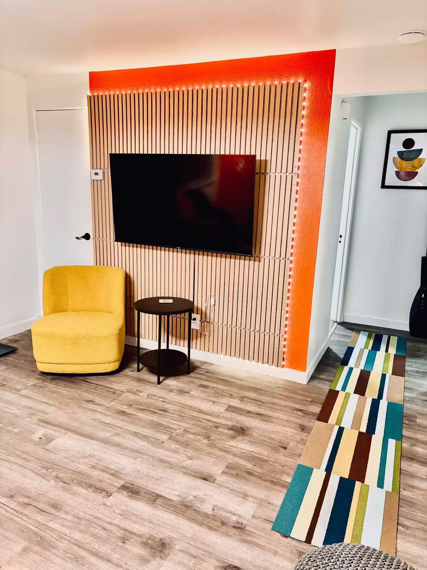 Un salón con un televisor de pantalla plana montado en una pared de madera, un sillón amarillo, una mesita redonda y un tapete colorido a rayas.