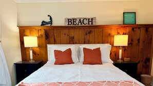 Hotelkamer met een houten hoofdbord, twee rode kussens, witte beddengoed en een bord met een strandthema boven het bed.