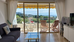 A balcony with a view of a cityscape and hills, a sofa, a glass table, and a TV on a cabinet. - Family apartment with swimming pool and sea view, quietly located with parking. (Cannes)