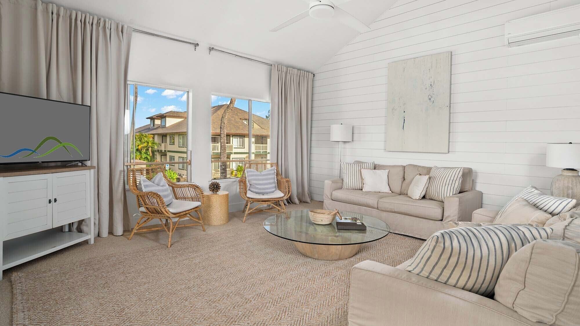 A spacious living room with a large TV, wicker chairs, a beige sofa, and a glass coffee table.