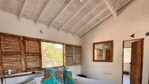 Un dormitorio con una cama grande, contraventanas de madera, un ventilador de techo y vistas a palmeras afuera.