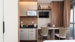 Cozinha compacta com micro-ondas, pia e armários de madeira. Há duas cadeiras de vime e uma TV fixada na parede.