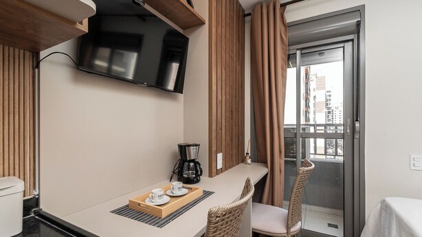 Quarto de hotel com cozinha compacta, cafeteira, televisão, cadeiras de vime e vista para a cidade através de uma janela grande.