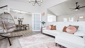 Ein geräumiges Wohnzimmer mit weißem Sofa, einem Hängekorbstuhl, einem flauschigen Teppich und einem Essbereich mit Tisch und Stühlen.