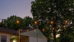 Un'area patio con un braciere, quattro sedie di legno e un piccolo capanno decorato con luci a stringa.