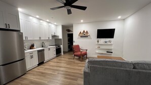 A modern kitchen with white cabinets, a stainless steel refrigerator, and a built-in oven. There is a grey sofa and a red chair in the living area, and a flat-screen TV mounted on the wall. - Brand New Modern Mountain Suite w/heated driveway and sidewalks (Revelstoke)