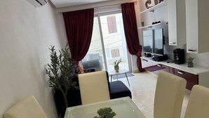 Ett modernt vardagsrum med ett matbord med glasskiva, beige stolar, en tv och en krukväxt.