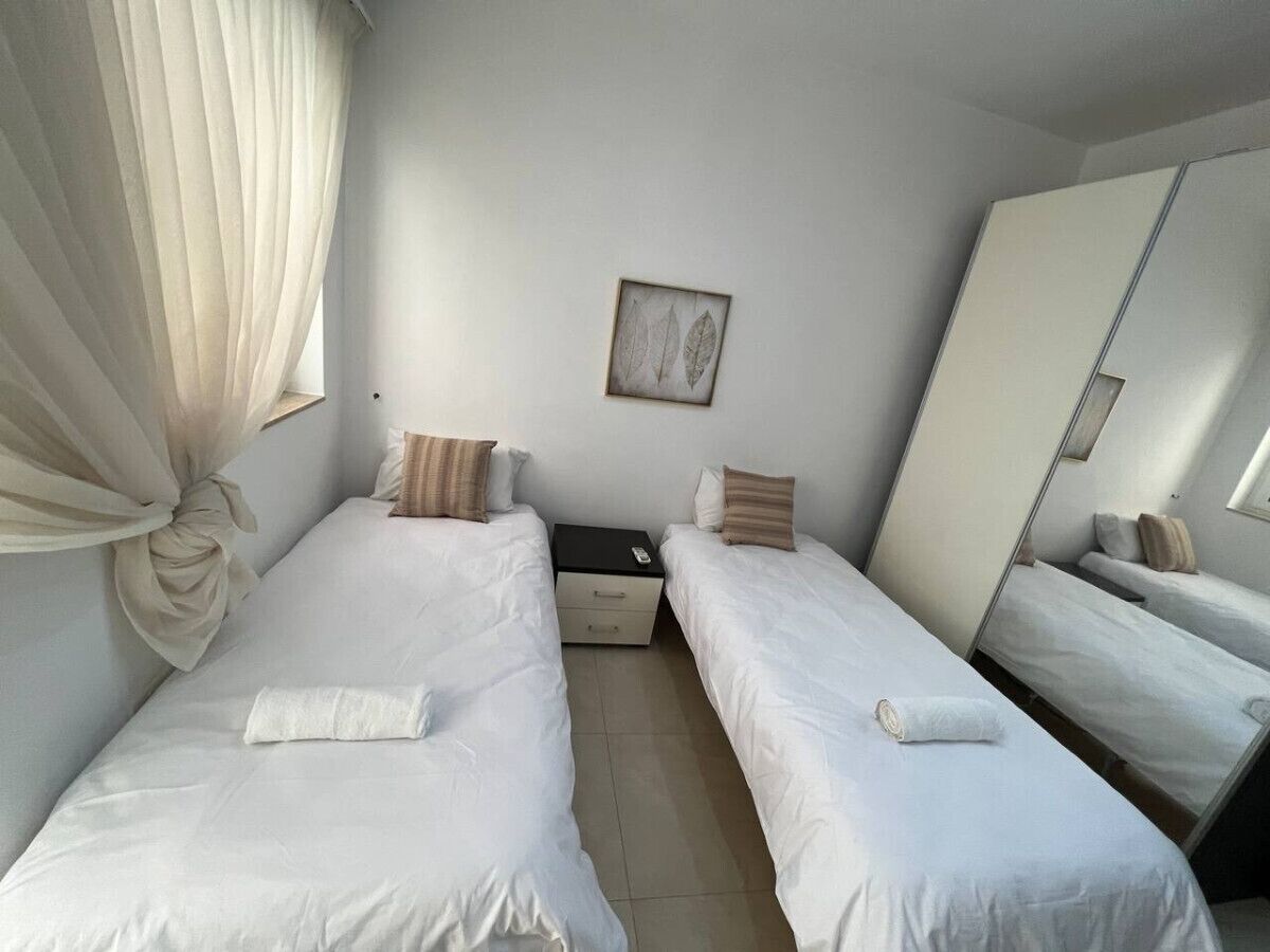 A hotel room with two single beds, a small bedside table, a nightstand, a mirror, and a window with sheer curtains.